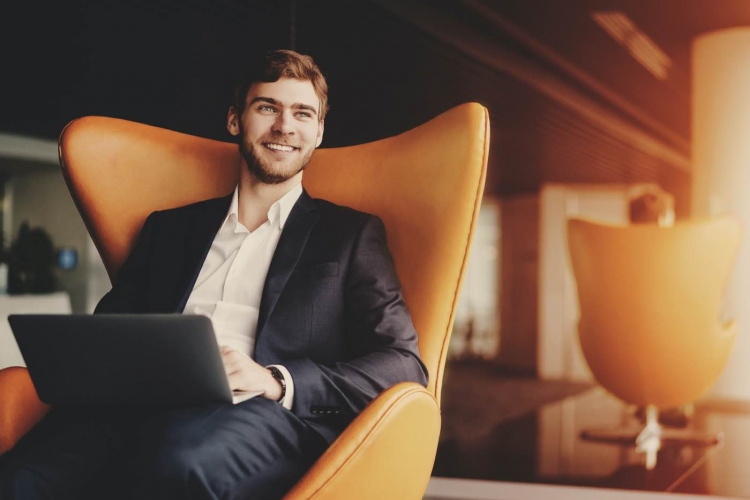 Joven empresario exitoso en la oficina de negocios sentado en un sillón naranja con ordenador portátil.