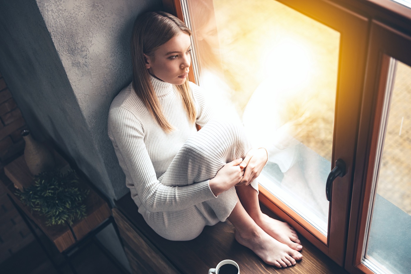 Mujer joven mirando por la ventana mientras bebe una taza de café.