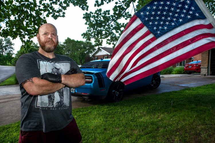 Hombre calvo con barba en el patio delantero de su casa, bandera estadounidense y muscle car