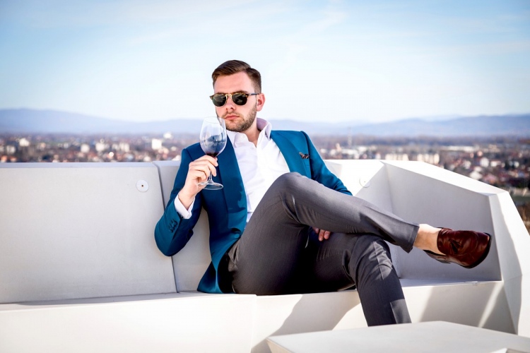 Hombre millonario con un caro traje hecho a medida, sentado al aire libre con gafas y sosteniendo una copa de vino tinto