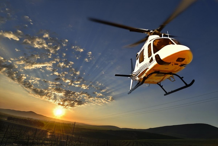 Helicóptero modelo Augusta volando sobre viñedos iluminados por el sol de la tarde.