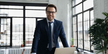 Hombre de negocios en gafas posando en su oficina moderna, en traje formal mira a cámara, muestra confianza en el éxito en el lugar de trabajo.