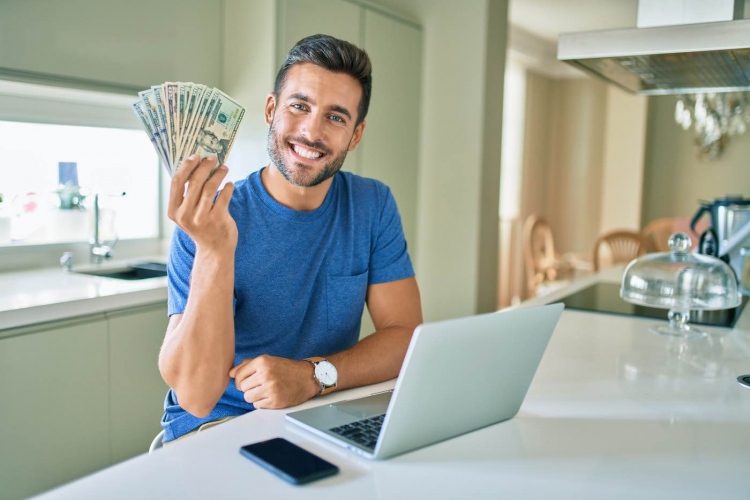 Hombre sostiene billetes de dólares americanos en casa frente una computadora portátil.