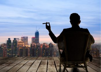 Multimillonario sentado en una silla, sostiene un cigarro y mira la ciudad en la noche.